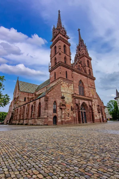 Basel 'deki tarihi Gotik katedralde ikiz kuleler, taş cephe, kaldırım taşı meydan ve dramatik gökyüzü.
