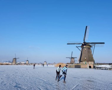 İnsanlar güneşli kış günlerinde Hollanda 'da birçok yel değirmeniyle anaokulu yakınlarında buzda kayıyorlar.