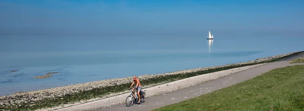 Férfi a kerékpár pályán és magányos vitorlás hajó hatalmas üres kék tó Zeeland — Stock Fotó