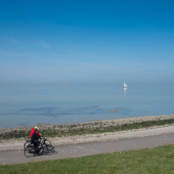 Mensen rijden op fietspad en eenzame zeilboot op uitgestrekte lege blauwe meer in zeeland — Stockfoto