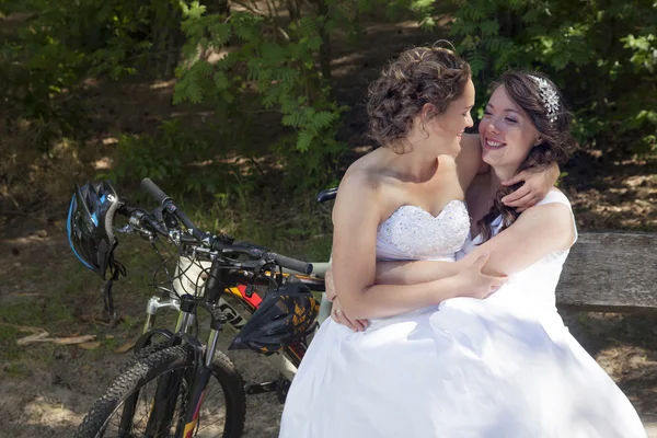 Two brides on bench in forest with mountain bikes — Stock Photo, Image