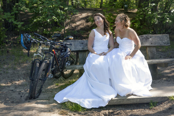 two brides on bench in forest with mountain bikes