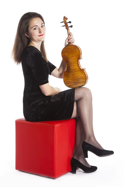 Morena com violino senta-se no banquinho vermelho no estúdio e sorri — Fotografia de Stock