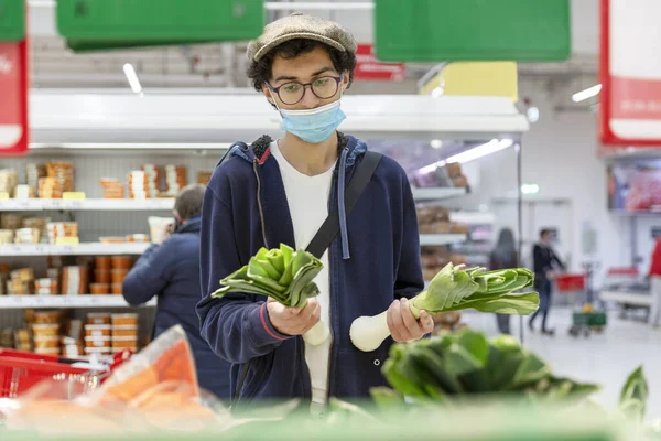 A young man wearing a medical mask picks a leek in the vegetable section of a supermarket. Healthy eating and vegetarianism. Coronavirus pandemic.