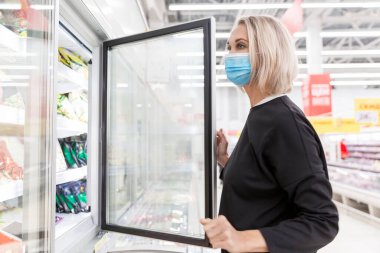 Donmuş gıda bölümünde, büyük bir süpermarkette tıbbi maske takan genç bir kadın. Koronavirüs salgını.