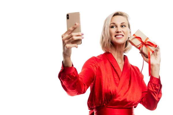 一个手里拿着圣诞礼物的笑着的年轻女人在电话里给自己拍照 漂亮的金发姑娘穿着红色毛衣 被白色背景隔离 案文的篇幅 — 图库照片