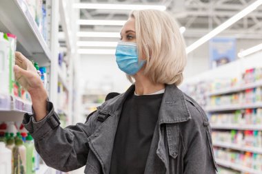 Süpermarkette tıbbi maskeli genç bir kadın temizlik ürünlerini seçiyor. Güzel sarışın. Koronavirüs salgını.
