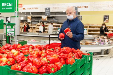 Süpermarketin sebze reyonunda maskeli bir adam elinde kırmızı dolmalık biber tutuyor. Moskova, Rusya, 09 / 15 / 2020.