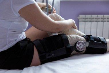 Bacak ameliyatından sonra ortoz ve elastik bandajlı bir kadın hastanede bir yatakta oturuyor..