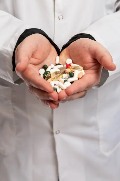A doctor in a dressing gown holds a handful of multicolored pills and capsules in his hands. Pharmacology and Medicine. Vertical.