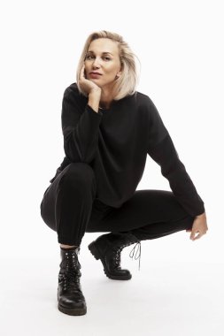 Siyah takım elbiseli, gülümseyen sarışın kadın çömeliyor. Beyaz arka plan. Dikey.