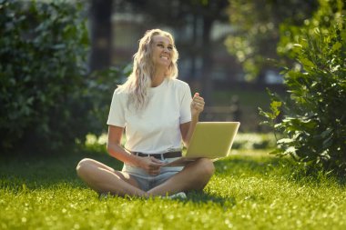 Dizüstü bilgisayarı olan genç bir kadın güneşli bir günde parkta çimlerin üzerinde oturur. Beyaz atletli ve şortlu, gülen bir sarışın. Çevrimiçi eğitim, uzaktan çalışma ve sosyal ağlarda iletişim.