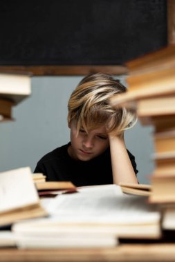 Siyah tişörtlü üzgün bir çocuk okul yönetim kurulunun arka planında bir sürü kitapla bir masada oturuyor. Salgın sırasında uzaktan eğitim. Gri arka plan. Mesaj için yer var. Dikey.