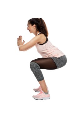 Spor kıyafetli gülümseyen genç bir kadın çömelme hareketi yapıyor. Spor, hareket ve aktif yaşam tarzı. Profil görünümü. Beyaz arka planda izole edilmiş. Dikey.