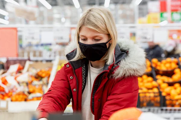 Young woman in a black medical mask in the supermarket in the department with fruits and vegetables. Quarantine during the coronavirus pandemic.