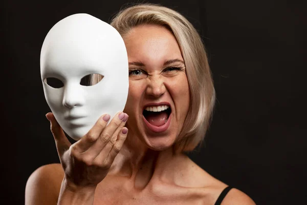 Screaming young blonde woman with a white mime mask in her hand. Change of emotions. Black background.