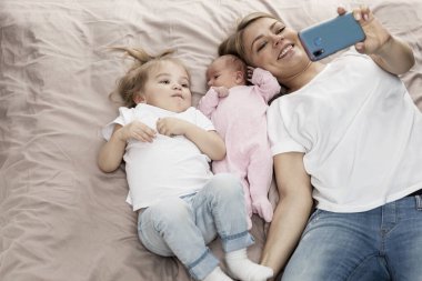 Anne ve küçük kızları yatağa uzanıp selfie çekiyorlar. Video oyunları ve çizgi film izlemek. Rahatlama ve aileyle iletişim.