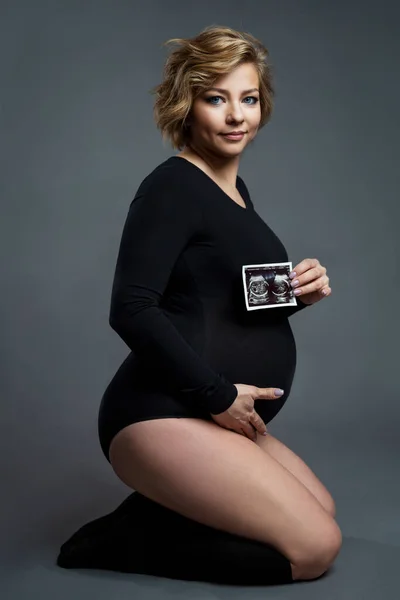 一位身穿黑色紧身衣的年轻漂亮的孕妇坐在那里 手里拿着超声波扫描 幸福就是等着孩子 灰色背景 垂直方向 — 图库照片