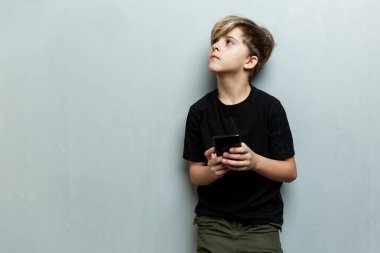 Elinde telefon olan bir çocuk ayağa kalkar ve bakar. Gri arka plan. Metin için boşluk.