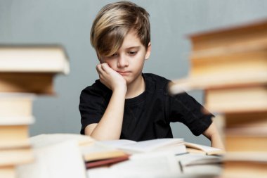 9-10 yaşlarında düşünceli bir çocuk kitaplarla dolu bir masada oturuyor. Gri arka plan. Sınavlar ve öğrenme sorunları.
