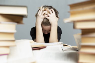9-10 yaşlarında, stresli bir çocuk kitaplarla dolu bir masada oturuyor ve ellerini başının arkasında paylaşıyor. Gri arka plan. Sınavlar ve öğrenme sorunları.