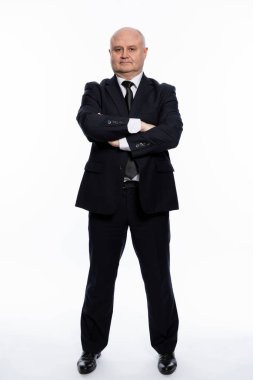 Kel, orta yaşlı, katı siyah takım elbiseli bir adam kolları göğsünde duruyor. İş ve şirket tarzı. Tam yükseklik. Beyaz arka plan. Dikey çizgi.