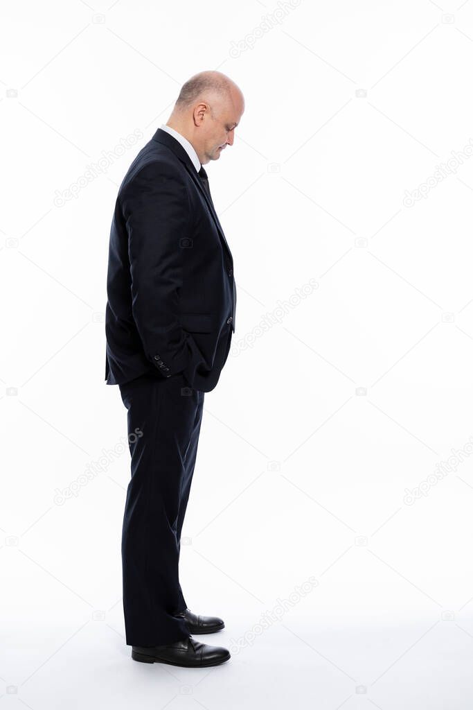 A bald middle-aged man in a strict black suit stands and looks down, holding his hands in his pockets. Business complexities. Side view. Full height. White background. Vertical.