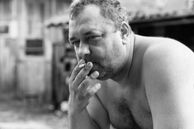 Çıplak gövdeli yetişkin bir adam köydeki bir evin avlusunda sigara içiyor. Depresyon ve stres. Siyah beyaz fotoğraf.