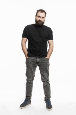 Sakallı bir adam elleri cebinde duruyor. Kot pantolonlu ve siyah tişörtlü genç bir adam. Tam yükseklik. Beyaz arka plan. Dikey.