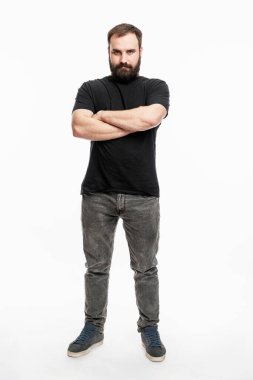 Sakallı bir adam ayakta, kolları göğsünde çapraz duruyor. Kot pantolonlu ve siyah tişörtlü genç bir adam. Tam yükseklik. Beyaz arka plan. Dikey.