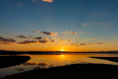 Tepeler ve göl üzerinde gün batımı. Gün batımında bulutların silueti. Günbatımı arkaplan fotoğrafı. Tepelerin yansımaları ve suyun üzerindeki bulutlar.