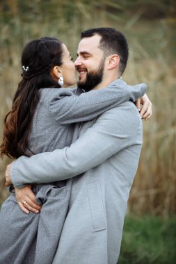 Gri ceketli şık bir çift arka planda nazikçe sarılıyor. Mutlu şehvetli düğün çifti kucaklaşması. Yeni evlilerin romantik anları. Dikey çerçevelenmiş çekim.