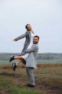 Doğanın arka planında gri ceketli şık bir çift. Damat gelini kaldırır. Düğün gününün mutlu anları. Dikey çerçevelenmiş çekim.