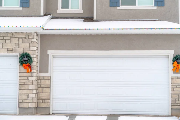 White garage door of home with wreath and snowy roof lined with christmas lights
