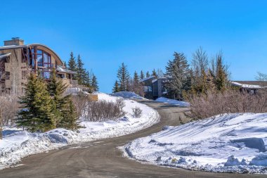 Karlı bir mahallede, kar ve ağaçların arasında evleri olan kıvrımlı asfalt bir yol.