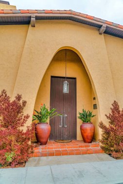 Kahverengi ahşap kapılı kemerli giriş. Etrafı bitkilerle çevrili büyük kavanozlarla çevrili.