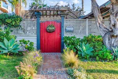 Beyaz çitin içinde patikası olan bahçe ve pergola ile kırmızı kapı.