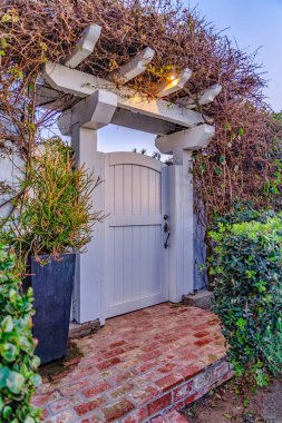 Pergola ve çitlerle kaplı beyaz ahşap kemerli kapı. Aşırı büyümüş sarmaşıklarla kaplı.