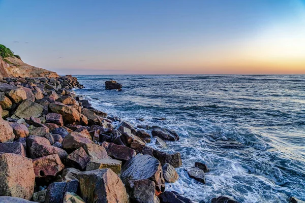 Havsvågor kraschar på ojämna klippor med blå himmel och gyllene sol vid solnedgången — Stockfoto