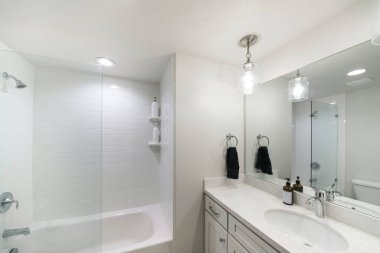 Küçük, modern, düz, beyaz bir banyo, cam duvar bölmesi ve beyaz fayansları olan bir küvet.