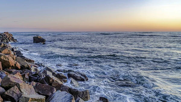 Pano Ocean vågor kraschar på ojämna stenar med blå himmel och gyllene sol vid solnedgången — Stockfoto