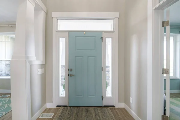 Mint front door interior with transom window and sidelights. Interior of a house with light gray walls, wooden flooring and a view of two rooms on both sides with windows.