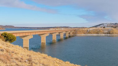 Pano kenarlarında kar ve kenarlarında köprü olan berrak ve huzurlu bir göl.