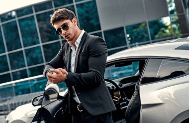 Takım elbiseli yakışıklı genç işadamı modern iş merkezinin önünde spor arabasının yanında duruyor.