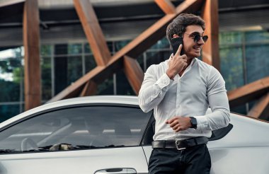Takım elbiseli yakışıklı genç işadamı modern iş merkezinin önünde spor arabasının yanında duruyor ve cep telefonuyla konuşuyor.