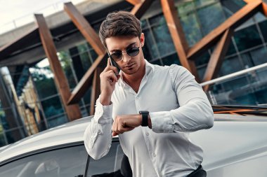 Takım elbiseli yakışıklı genç işadamı modern iş merkezinin önünde spor arabasının yanında duruyor.