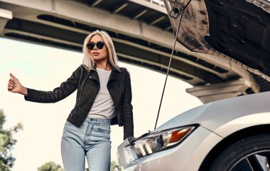 Sokakta kaputu açık, kırık bir arabanın yanında duran günlük kıyafetleri ve güneş gözlükleriyle çekici genç bir sarışın..