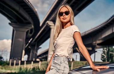 Sokakta spor arabanın yanında duran günlük kıyafetleri ve güneş gözlükleriyle çekici genç bir sarışın. Şehir tarzı.