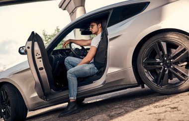 Modern spor arabasında oturan güneş gözlüklü ve günlük kıyafetli yakışıklı bir genç.