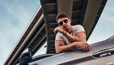 Sokakta modern spor arabasının yanında duran, günlük kıyafetli yakışıklı genç adam.
